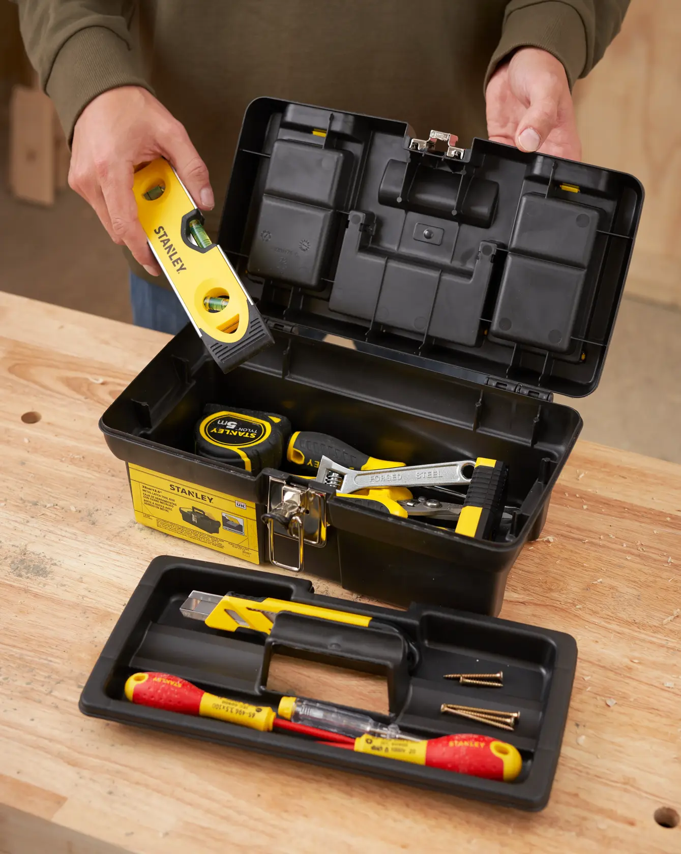 STANLEY 1-92-064 black plastic toolbox with tools and removable tray on workbench, person holding yellow level.