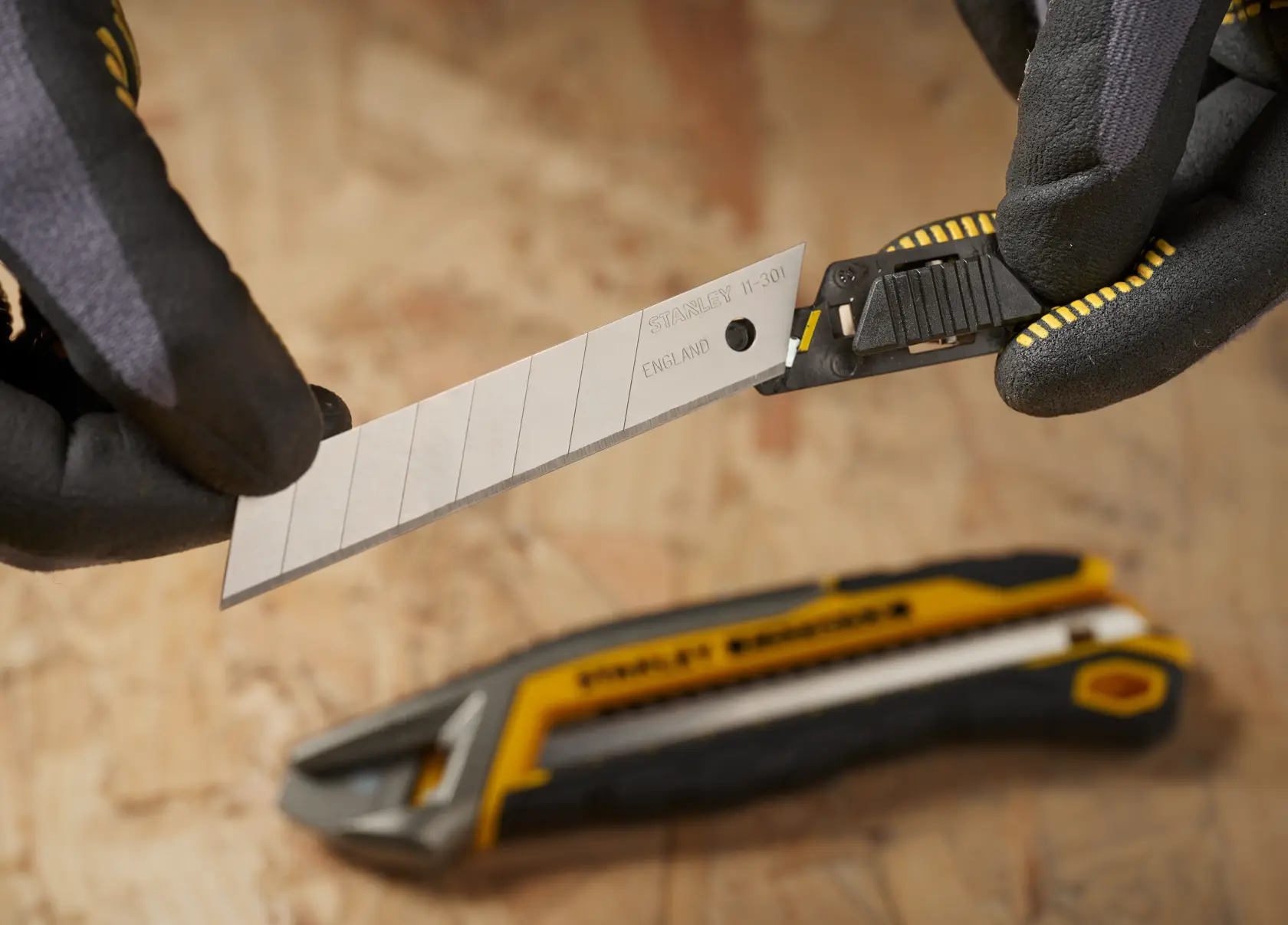 STANLEY 2-11-301 snap-off utility knife blade held in gloved hands, with utility knife handle on wood surface.