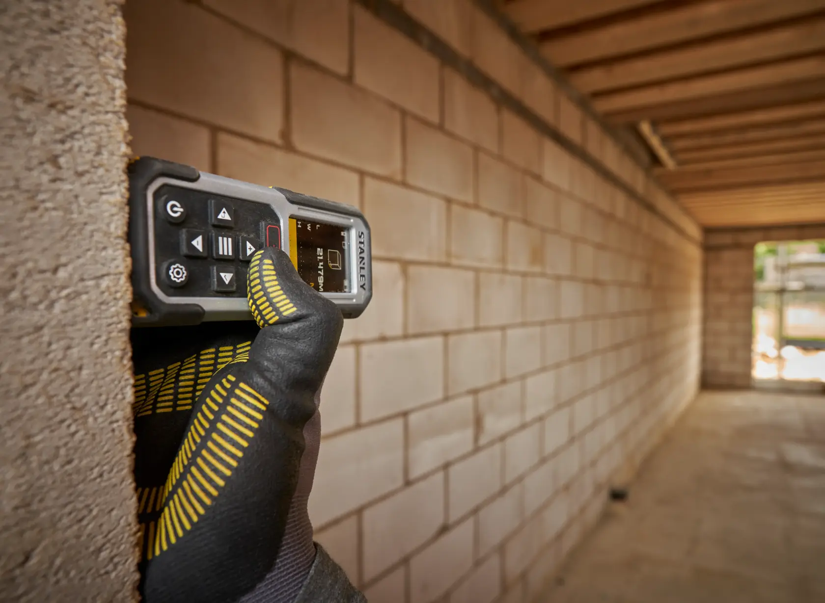 STANLEY FMHT77050-XJ laser distance measurer held by gloved hand against brick wall in construction site.