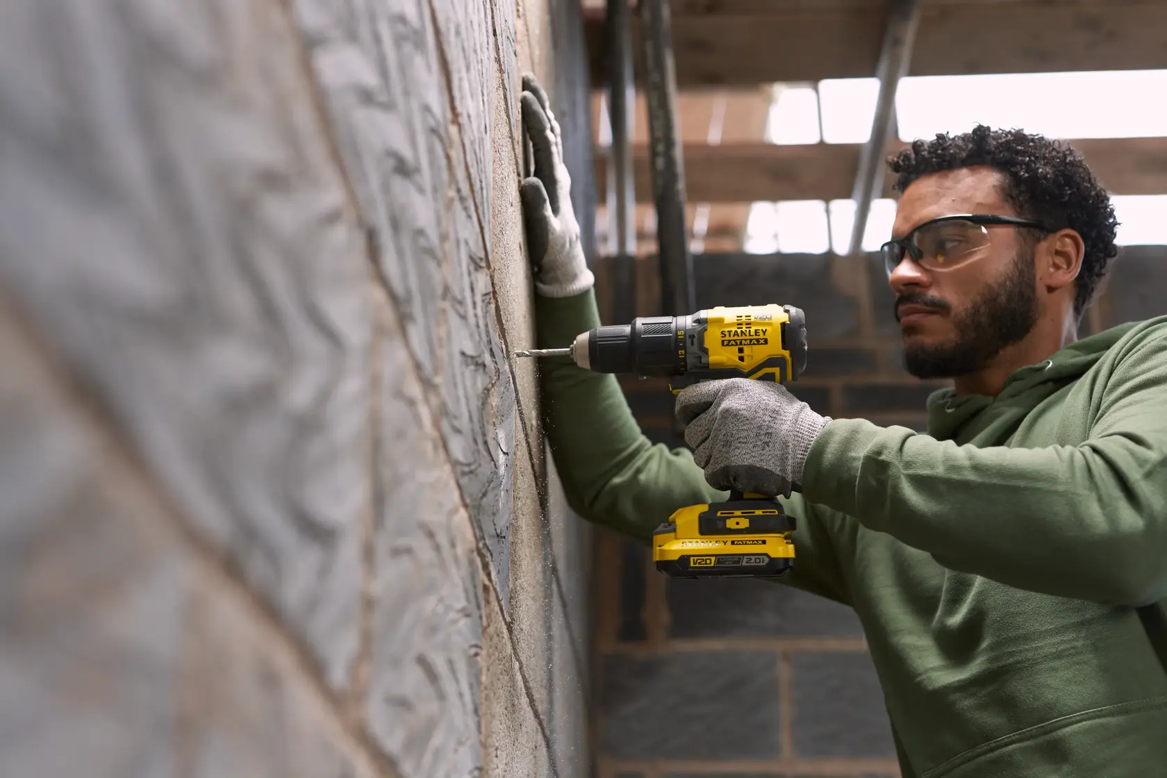 Person using STANLEY SFMCD715D1K cordless drill on a brick wall, wearing gloves, indoor construction setting.