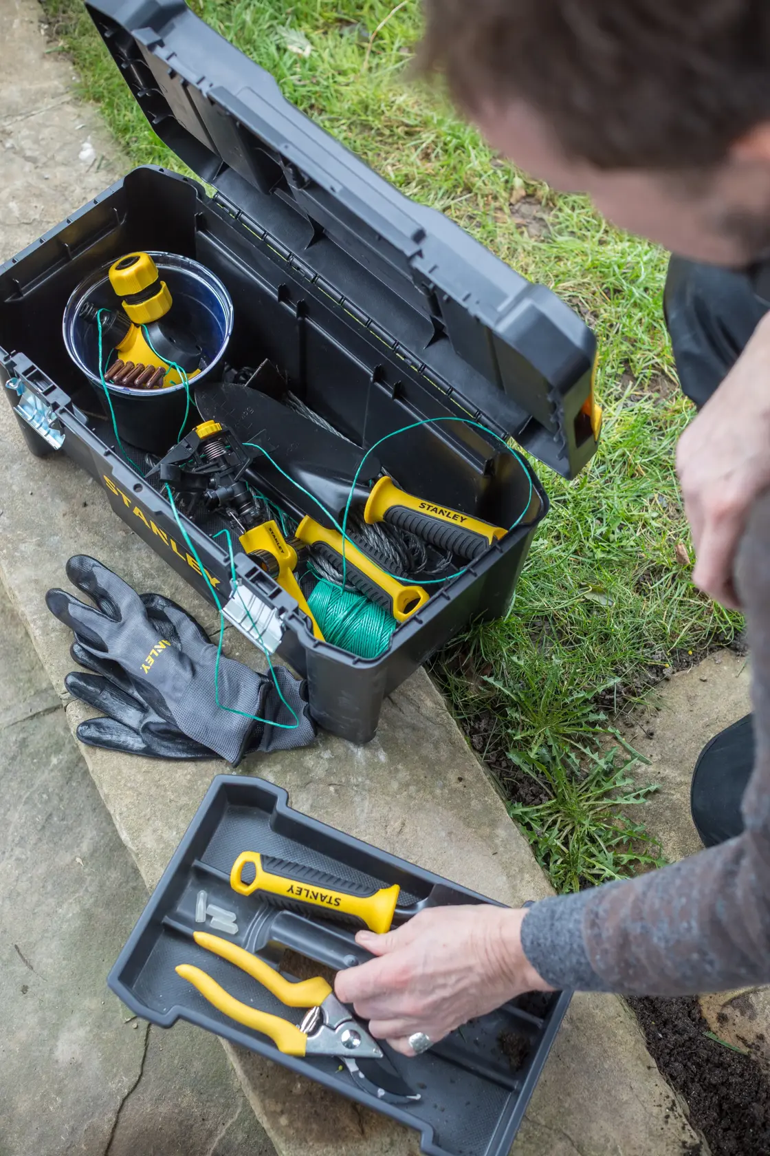 STANLEY STST1-75521 tool box with open lid, showing garden tools and gloves, top-down outdoor view.